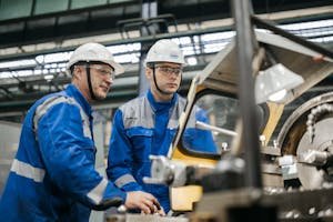 Two industrial workers in protective gear operate machinery in a factory setting.